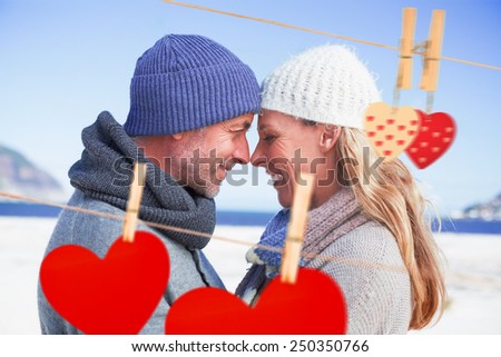 Attractive couple smiling at each other on the beach in warm clothing against hearts hanging on the line