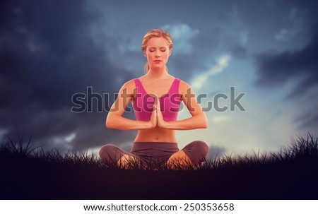 Calm blonde sitting in lotus pose with hands together against blue sky over grass