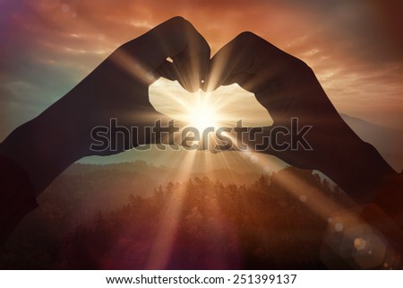 Woman making heart shape with hands against sunrise over mountains