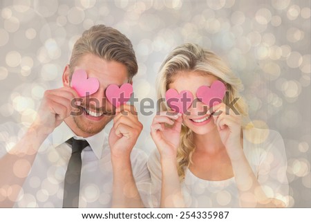 Attractive young couple holding pink hearts over eyes against light glowing dots design pattern