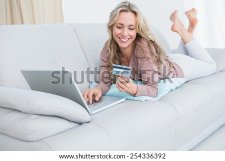 Smiling blonde lying on couch shopping online at home in the living room