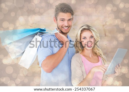 Attractive young couple holding shopping bags using tablet pc against light glowing dots design pattern