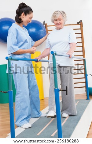 Senior woman walking with parallel bars with therapist in fitness studio