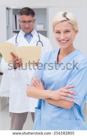 Portrait of confident nurse standing arms crossed while doctor reading reports in background at clinic
