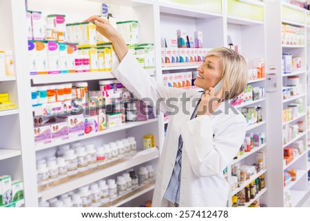 Smiling pharmacist phoning and taking medicine from shelf in the pharmacy