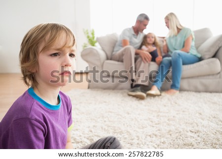 Upset boy sitting on floor while parents enjoying with sister on sofa at home