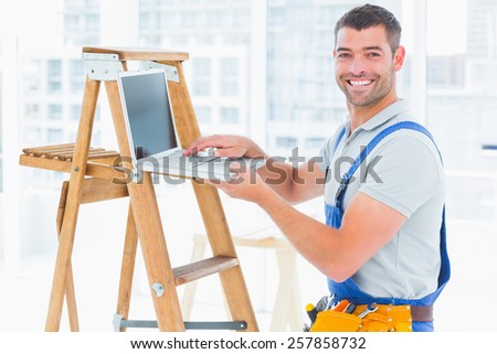 Portrait of smiling handyman using laptop by ladder in bright office