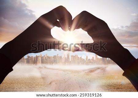 Woman making heart shape with hands against path in yellow field leading to city