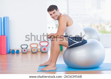 Smiling man sitting on exercise ball and looking at camera in fitness studio