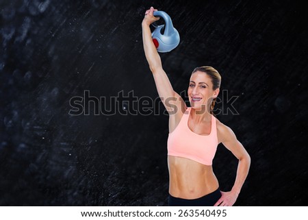 Female blonde crossfitter lifting kettlebell above head smiling at camera against black background