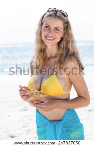Smiling pretty blonde in bikini holding a starfish and looking at camera at the beach