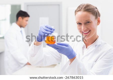 Smiling scientist looking at camera and holding beaker with orange fluid in laboratory