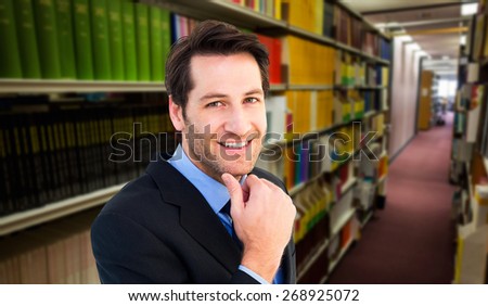 Businessman touching his chin while smiling at camera against rows of bookshelves in the library