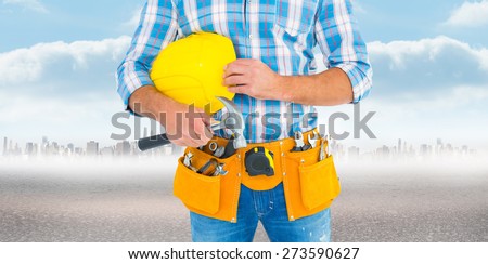 Manual worker wearing tool belt while holding hammer and helmet against city on the horizon