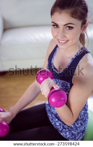 Fit woman lifting dumbbells on exercise ball at home in the living room