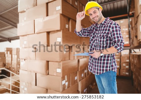 Happy repairman wearing hard hat while holding clipboard against shelves with boxes in warehouse