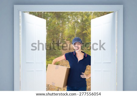 Happy delivery woman holding cardboard box against scenic view of walkway along lush forest