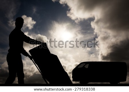 Happy delivery man pushing trolley of cardboard boxes against dark sky with white clouds