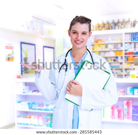 Doctor holding clipboard and presenting with her hand against close up of shelves of drugs
