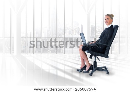 Businesswoman sitting on swivel chair with laptop against room with large window looking on city