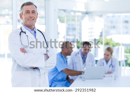 Happy doctor looking at camera with arms crossed while his colleagues works in medical office