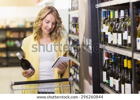 Smiling pretty blonde woman having a wine bottle in her hand and looking at a notepad in supermarket