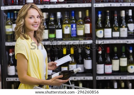Portrait of a smiling pretyt blonde woman having in her hands a wine bottle and notepad in supermarket