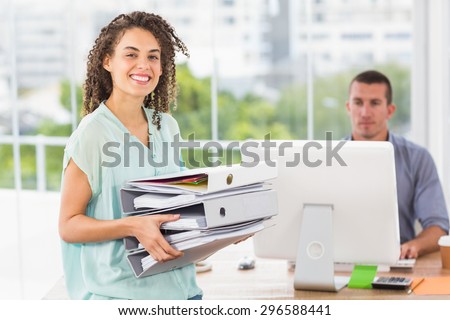 Smiling businesswoman carrying a stack of folders while her colleague typing on computer