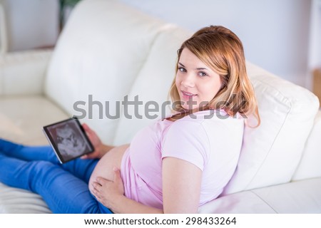 Pregnant woman looking at camera with hand on belly and ultrasound scans in the living room