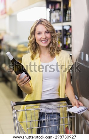 Portrait pf a smiling pretyt blonde woman having a notepad and wine bottle in her hands in supermarket