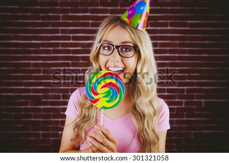 A beautiful hipster with party hat holding a giant lollipop against a red brick wall