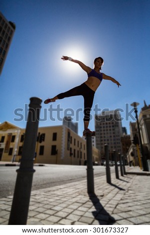 Athletic woman balancing on bollard and stretching out her arms and leg in the city