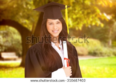A woman standing to the side slightly with her degree and dressed in her graduation robe against trees and meadow in the park
