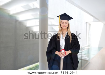 Blonde student in graduate robe holding her diploma against stylish modern home interior with staircase