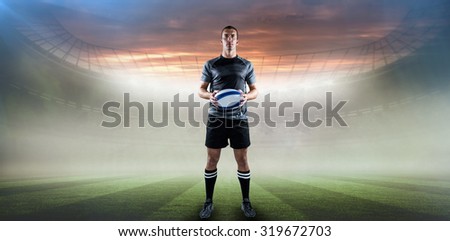 Serious rugby player in black jersey holding ball against rugby pitch
