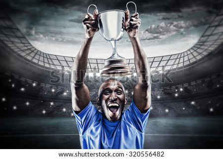 Happy athlete cheering while holding trophy against large football stadium under blue sky