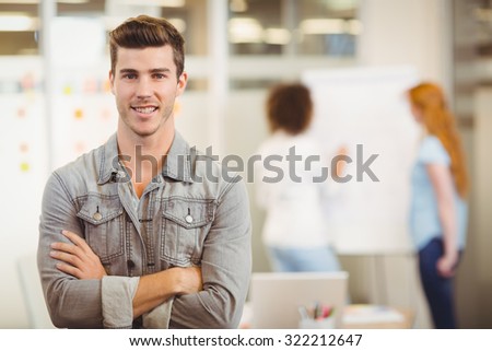 Portrait of smiling businessman with arms crossed standing against female colleagues working office