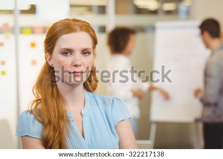 Portrait of serious businesswoman with arm crossed sitting on table with colleagues looking at whiteboard in background at office