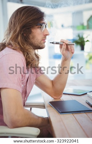 Side view of hipster smoking electronic cigarette at desk in office