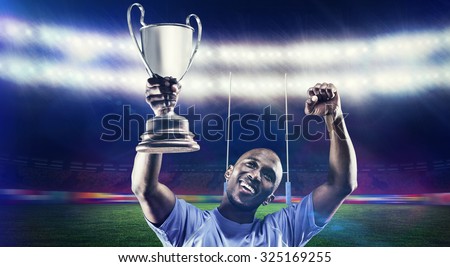 Happy sportsman looking up and cheering while holding trophy against rugby stadium