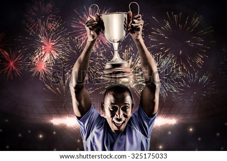 Portrait of happy athlete cheering while holding trophy against fireworks exploding over football stadium