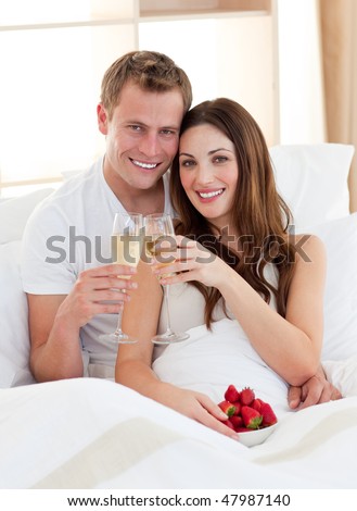 Enamoured couple drinking champagne with strawberries lying in bed at home