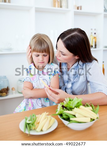 Dissatisfied blond girl eating vegetables with her mother in the kitchen