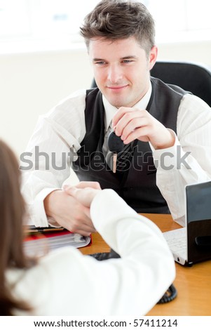 Young attractive salesman giving car's key to his customer sitting in his office