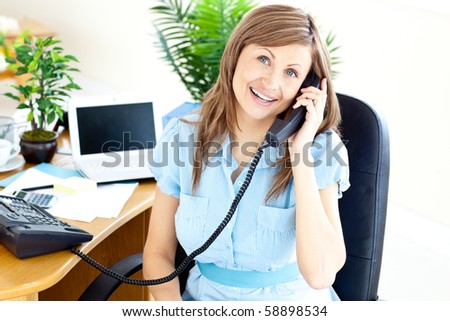 Lively young businesswoman talking on phone sitting at her desk in the office