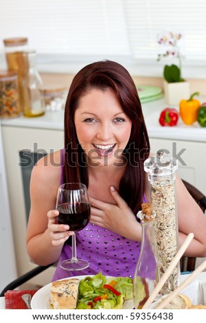 Cheerful woman eating her meal holding a glass of wine at home in the kitchen