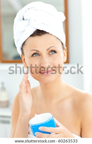 Positive young woman with a towel putting cream on her face in the bathroom at home