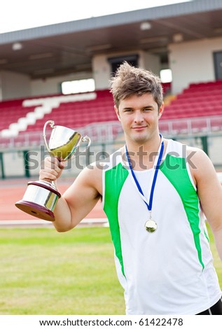 Delighted male athlete holding a cup and a medal standing in a stadium