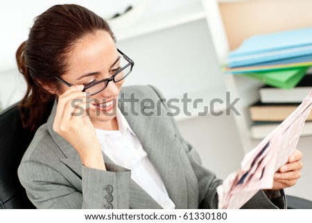 Cheerful businesswoman holding her glasses and reading a newspaper in her office sitting at her desk