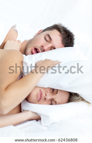 Scenic in bedroom of young couple lying in bed and holding pillow on the ears because of snoring boyfriend.